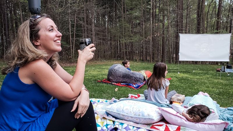 A mom and her three children sit outside on blankets and pillows. There is a white screen set up in the yard and trees all around. The mom is holding a stemless glass of wine.